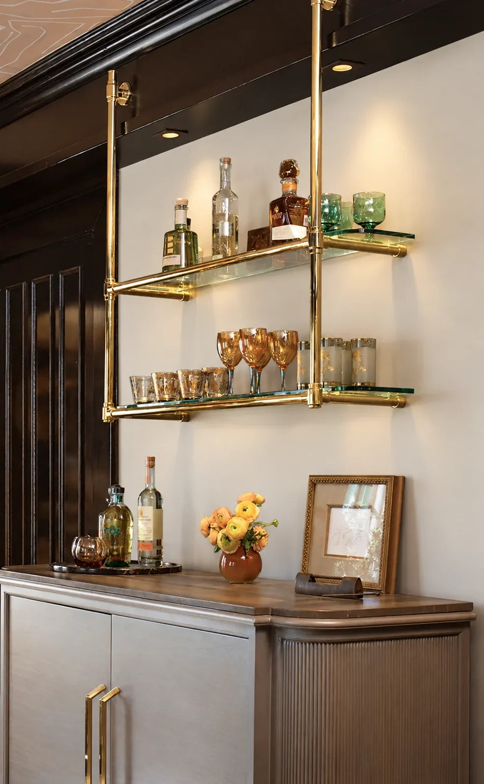 A modern wall-mounted double glass shelf with gold metal framing, styled with bottles and glasses in an elegant interior setting.