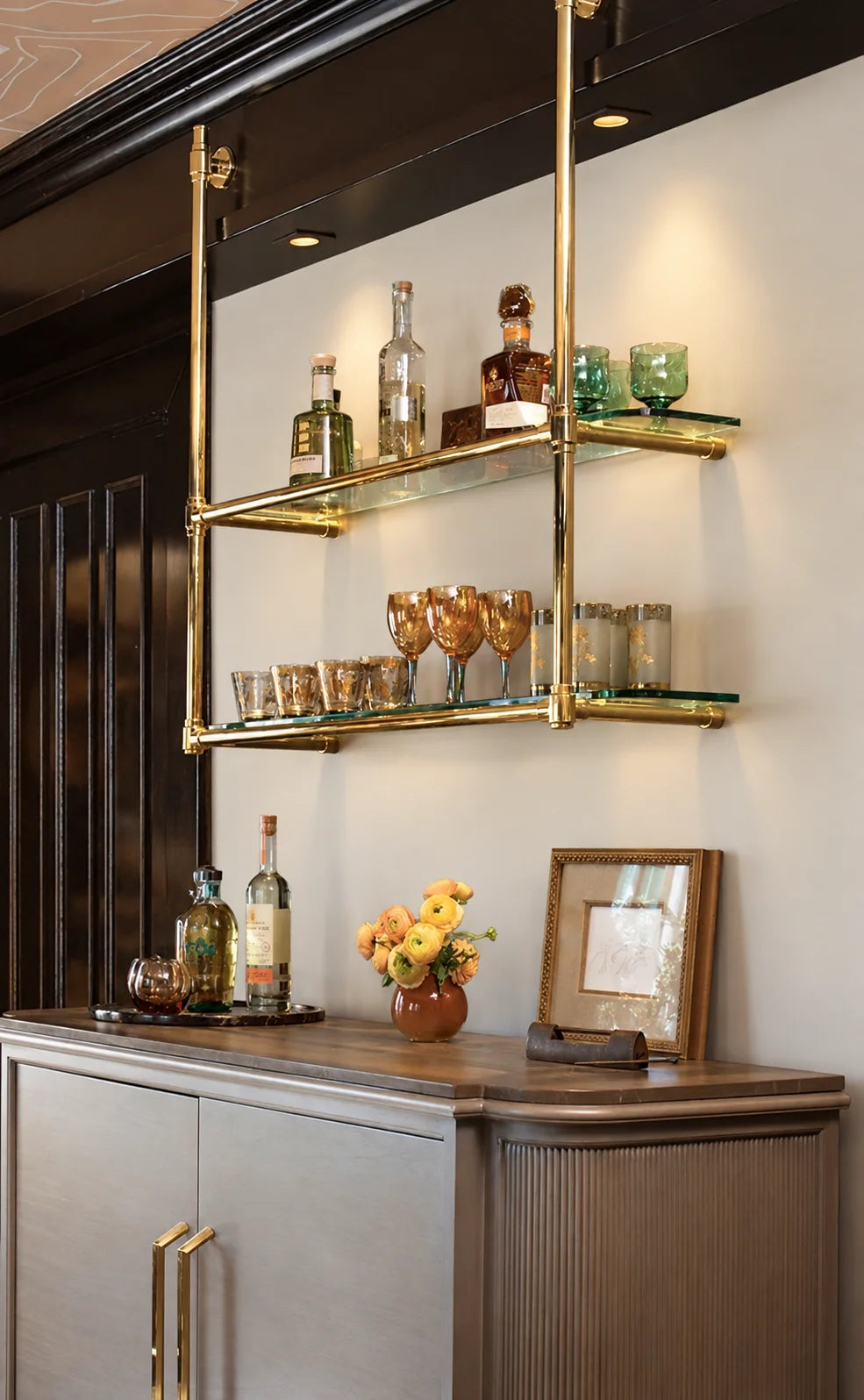 A modern wall-mounted double glass shelf with gold metal framing, styled with bottles and glasses in an elegant interior setting.