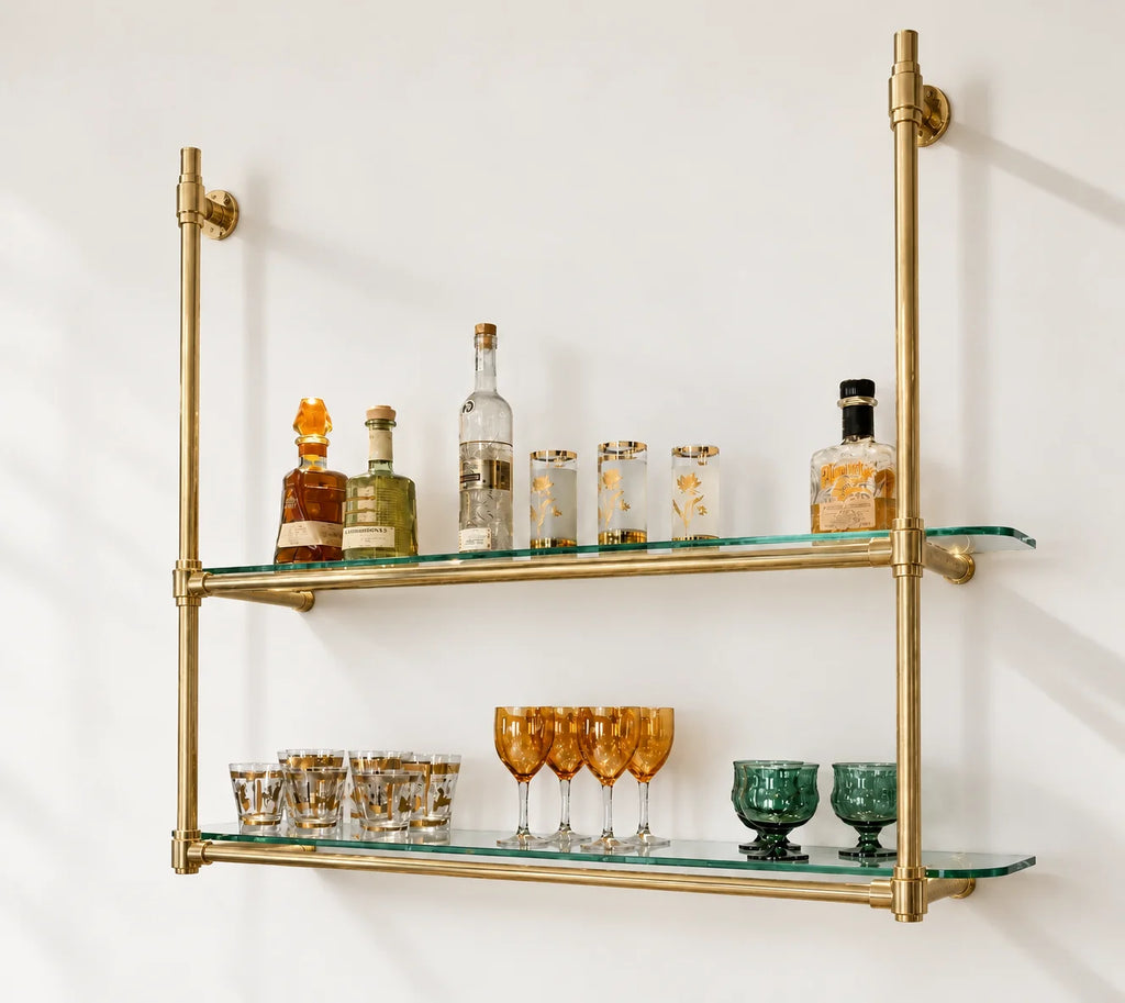 Industrial-style double glass shelf mounted on a white wall, made of brass pipes with clear tempered glass shelves, styled with bottles, glasses, and tumblers.