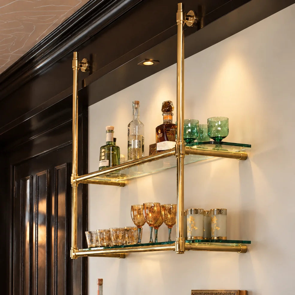 A wall-mounted brass bar shelving unit with two glass shelves, styled with bottles, glasses, and a small vase of flowers above a wooden cabinet in an elegant interior.