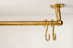 A polished brass hanging rod with two hooks, mounted to a ceiling with a matching ceiling mount, shown against a plain white wall.