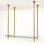 A modern brass wall-mounted shelf with four vertical hanging rods and a clear glass shelf, shown against a clean white wall and ceiling.
