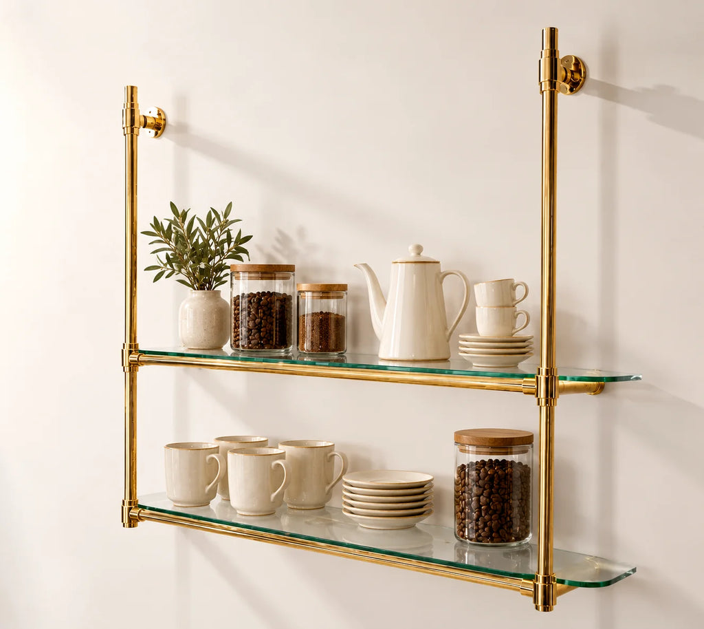 Two-tier wall-mounted glass shelves with a polished brass frame, styled with ceramic mugs, a teapot, a small plant, and glass canisters on a warm off-white wall.