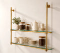 Two-tier wall-mounted glass shelves with a polished brass frame, styled with ceramic mugs, a teapot, a small plant, and glass canisters on a warm off-white wall.