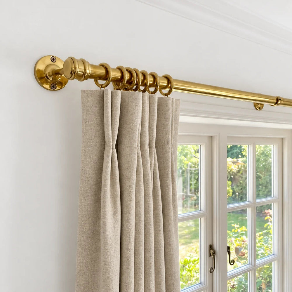A brass curtain rod with matching rings mounted on a white wall above a window, styled with beige linen curtains.