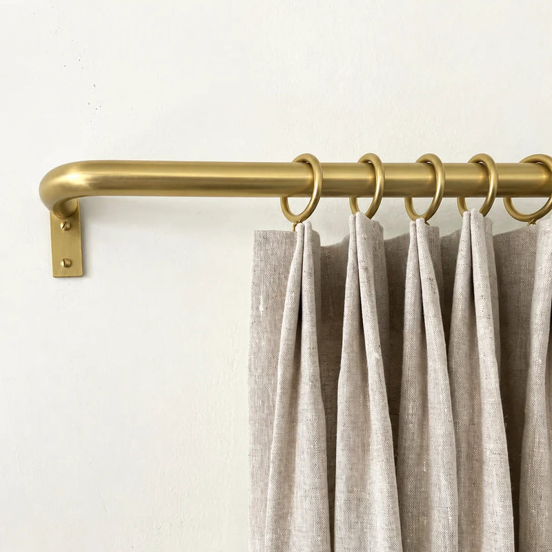 A close-up of light beige pleated curtains hanging from a brass curtain rod with matching rings against a plain off-white wall.