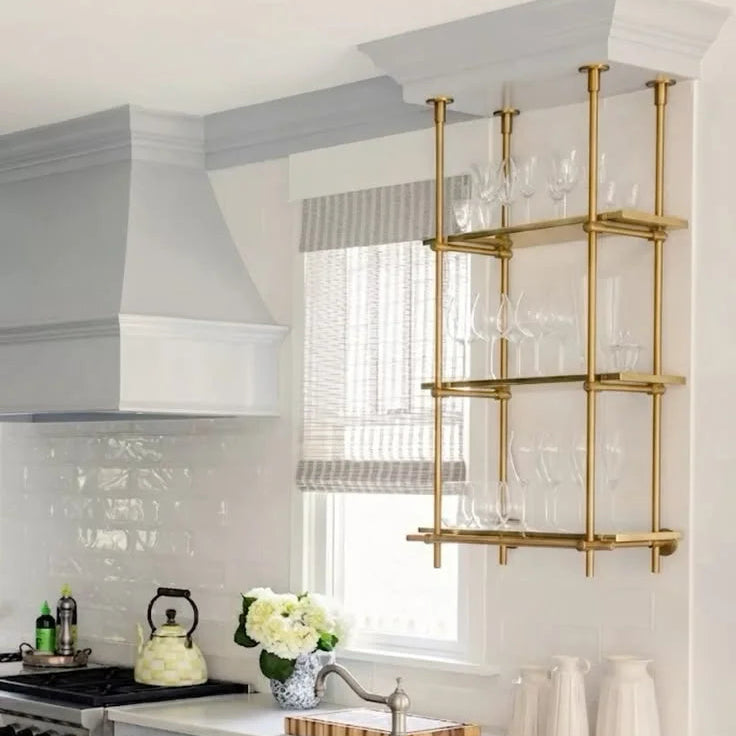 A modern kitchen interior with white cabinetry, a white countertop, a window with blinds, and a wall-mounted brass shelving unit holding clear glassware.