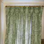 A pair of sage green damask curtains hanging from a gold curtain rod, shown in a home setting with white walls and molding.